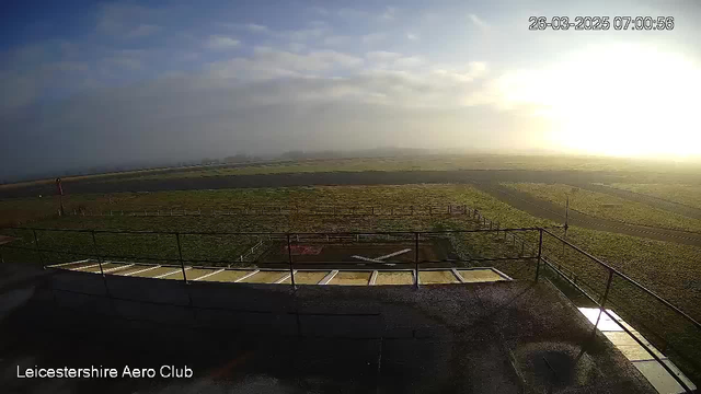 A panoramic view from a camera at the Leicestershire Aero Club, showing a grassy field and an airstrip under a cloudy sky. The sun is rising in the background, casting a warm glow, while a light fog covers part of the scene. In the foreground, there is a railing with a wooden platform and markings indicating a helipad area. The landscape is mostly flat with an airstrip visible in the distance.