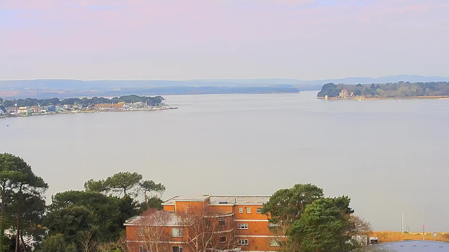 A view of a calm body of water stretching across the image, bordered by trees and buildings. On the left, there are several colorful structures along the shoreline, alongside some boats. In the distance, a small harbor and a wooded area can be seen. On the right side, a prominent building sits on a peninsula, surrounded by lush greenery. The sky above is light, suggesting a clear day.