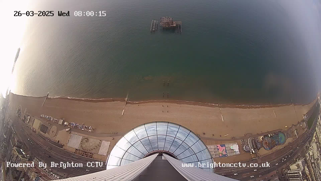 Aerial view of a beach and the sea taken from a high vantage point. The foreground shows a large circular glass structure at the top of a tower. Below, the sandy beach curves along the shoreline with a few wooden piers extending into the water. To the right, various colorful structures including amusement rides and a pool are visible along the beachfront. The sea appears calm and stretches out towards the horizon, where the sun reflects off the water, creating a bright, shimmering effect. The date and time are displayed in the upper left corner.