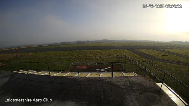 A view from a webcam at Leicestershire Aero Club showing an expansive green field and a runway. In the foreground, there is a ledge with railings, and an area marked with a red square containing a white "X" symbol, indicating a helicopter landing zone. The background features a foggy atmosphere with distant trees and rolling hills partially obscured by mist. The image shows an early morning setting with soft light.
