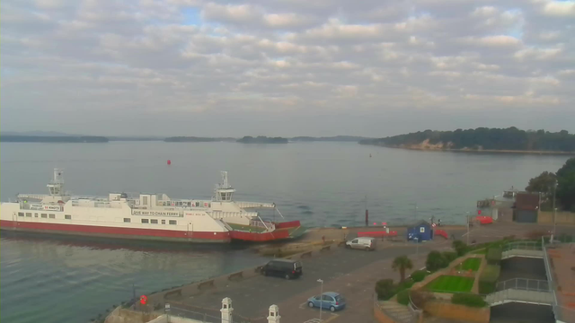 A ferry is docked at a quiet harbor on a calm body of water. The sky is partly cloudy with a mix of blue and gray tones. Nearby, there are a few vehicles parked and a path leading up to a landscaped area with palm trees and grass. In the distance, small islands can be seen, and a few buoys float in the water. The scene is tranquil, capturing a serene moment by the water's edge.