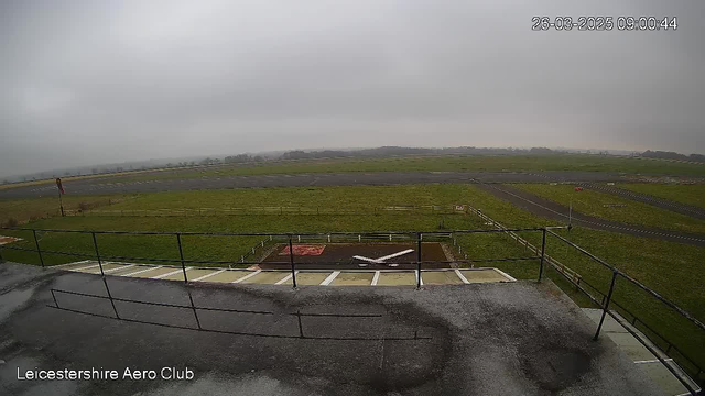 A view from a webcam showing an airfield on a cloudy day. The foreground features a fenced area with a white airplane silhouette on a gray surface. In the background, the flat grass runway stretches out, disappearing into a misty horizon. The sky is overcast, and the environment appears tranquil with no visible aircraft in the sky.