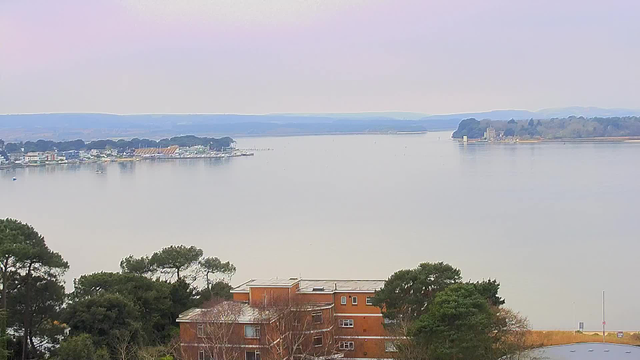A panoramic view of a calm lake or river, with distant hills visible in the background. On the left, there is a small marina with several boats and colorful buildings lining the shore. In the foreground, a cluster of trees and a red-brown building can be seen. The sky is lightly colored, suggesting early morning or late afternoon light, and the overall atmosphere is tranquil and serene.