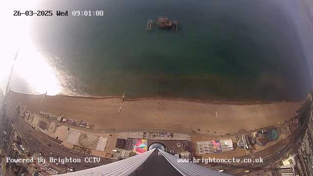 A high-angle view of a beach and ocean, taken from a webcam. The image shows a sandy shoreline with a few people walking along the beach. In the water, there are some boats, and a pier structure is visible further out in the ocean. The scene is bathed in bright sunlight, creating glimmers on the water's surface. On the right side of the image, there is a boardwalk with various amusement features and structures. The date and time are displayed in the upper left corner.