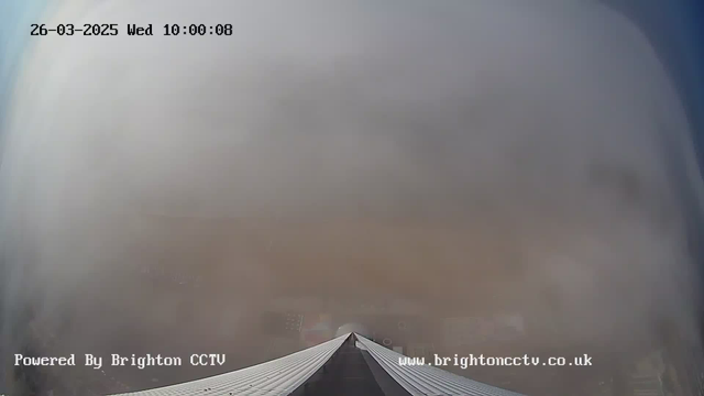 A view from a webcam showing a thick layer of fog or cloud, obscuring most of the landscape. The camera is positioned looking down a sloped roof, with the edges of the roof visible at the bottom of the image. The sky appears pale, and there are no identifiable features or colors visible in the background due to the fog. The timestamp in the upper left indicates the date and time as March 26, 2025, at 10:00:08. The image contains a watermark at the bottom left stating it is powered by Brighton CCTV.
