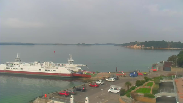 A large white ferry with a red stripe is docked at a pier surrounded by calm water. The background features a low, wooded shoreline. Several cars are parked near the dock, and there are some people on the ferry and around the pier. The sky is overcast, giving the scene a muted light.