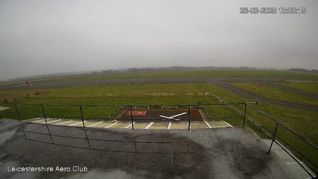 A cloudy and overcast scene at the Leicestershire Aero Club. In the foreground, there is a flat rooftop with a railing and a muddy area marked with a white cross. The background features a vast field of green grass alongside a gray asphalt runway. A distant horizon is visible, shrouded in mist, suggesting limited visibility. There is a flag on a pole gently fluttering in the wind.