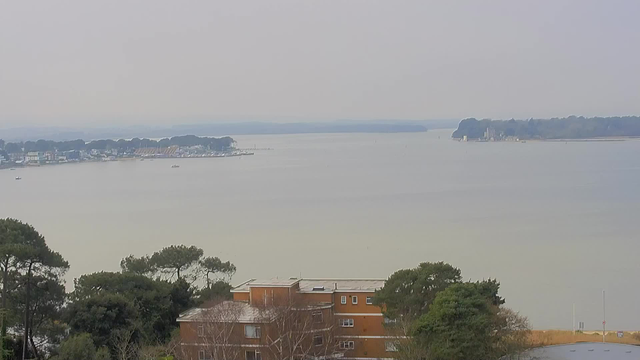 A serene view of a body of water stretching to the horizon, with a mixture of light gray and blue hues in the sky. In the foreground, there are several buildings, primarily in shades of orange and brown, along with trees. Toward the middle of the scene, boats are scattered on the water, and there are distant land formations and structures along the shoreline. The atmosphere appears calm and slightly hazy.