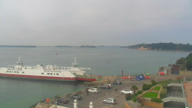 A view of a harbor with a large ferry docked, featuring a white and red color scheme. In the foreground, several parked cars are visible along a paved area. There are also a few people, some on bicycles, moving about near the water's edge. In the background, a calm waterway stretches out with several small islands visible in the distance. The sky is overcast, contributing to a soft, muted atmosphere. The shoreline has patches of greenery and well-maintained gardens.