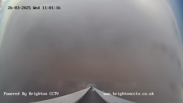 A webcam image showing a foggy view, where visibility is very low. The foreground features a rooftop with a sloped design leading to a point in the center. The background is mostly obscured by thick fog, blending the sky and landscape into a uniform grayish haze. The timestamp on the image reads 26-03-2025, 11:01:16.
