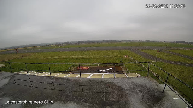 A view from a webcam at an airfield, with a cloudy sky above and an expansive green field below. In the foreground, there is a railing and a concrete area with a white airplane silhouette on the ground. The background features a runway with several sections of grass and a small portion of roadway visible. The image is timestamped, indicating the date and time.