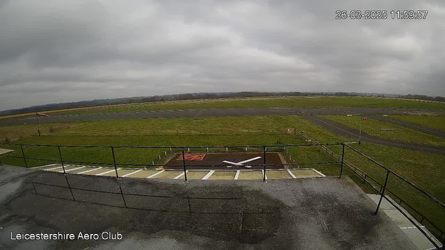 A view from a webcam showing an open grass field with a small runway in the background. The sky is overcast with gray clouds. A few markers and barriers are visible on the ground, with a small white airplane resting on a landing area. The area is surrounded by a railing, and there are signs of an aeroclub in the lower left corner of the image. The scene conveys a calm, cloudy day at an airfield.