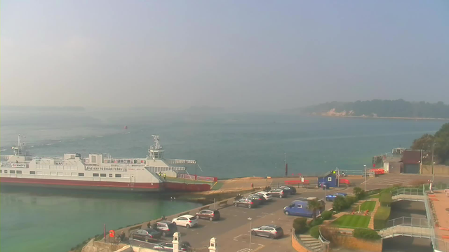 A view of a harbor with a large ferry docked near the shore. The ferry is white with red accents and has several windows. In the foreground, there are parked cars in a lot, with various vehicles visible. The background features a calm body of water that fades into a hazy horizon, where land can be seen in the distance. A few scattered trees are also visible along the shoreline. The sky is light blue with some haze, creating a soft and muted atmosphere.