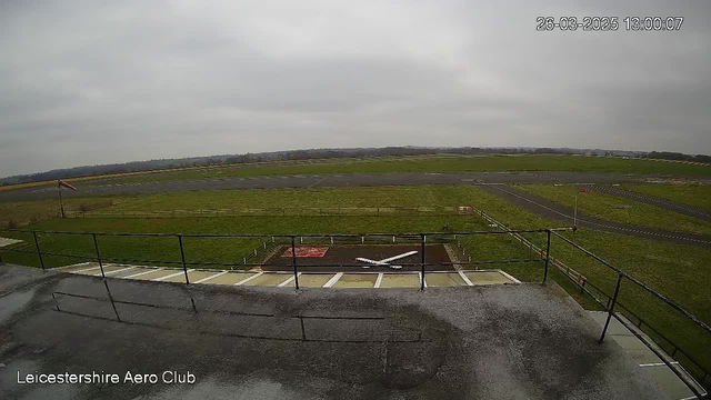 A view from a webcam at the Leicestershire Aero Club. The image captures an open green field with a tarmac runway in the background. A white cross-shaped glider is visible on a ground marking in the foreground. There are several fences around the area, and a flagpole with a flag waving gently in the breeze. The sky is overcast with a grayish tone, indicating cloudy weather.