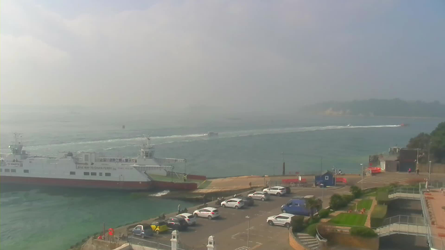 A view of a harbor with a large white and red ferry on the water, moving through a hazy atmosphere. In the foreground, rows of parked cars are visible along the shore. A smaller boat, creating ripples in the water, is making its way past the ferry. The shoreline features a few buildings, trees, and landscaped areas, while the distant background shows land fading into mist. The sky appears overcast with a soft light.