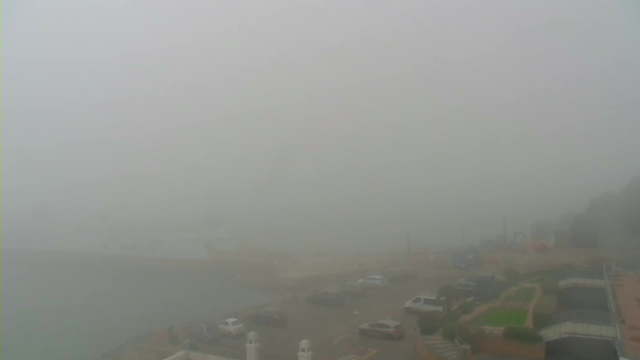 A heavily foggy scene showing a harbor area. The view is obscured, making it difficult to see details. In the foreground, there are several cars parked along a road. There is a hint of water and some structures in the background, but they are largely indistinct due to the fog. The overall atmosphere feels muted and gray.