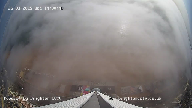 A webcam view looking down from a tall building, with a heavy layer of fog obscuring most of the landscape below. The only visible parts include the roofline of the building and some colorful shapes suggesting structures or artwork on the ground. The scene is primarily dominated by gray and white fog, creating a soft, indistinct atmosphere. The time and date are shown in the top left corner, along with a watermark for Brighton CCTV at the bottom.