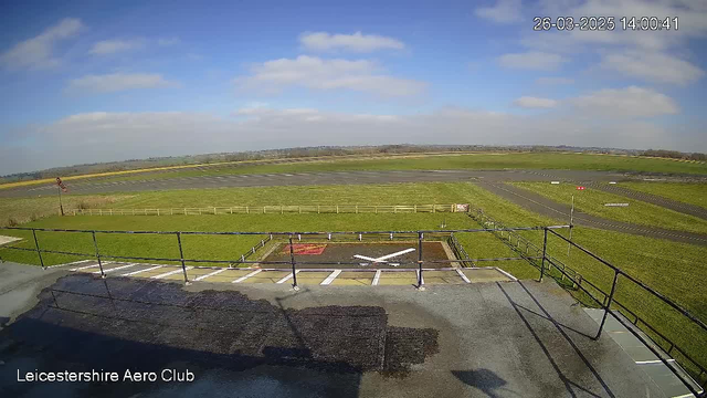 A clear view of an airport runway area taken from a high vantage point. In the foreground, there is a helicopter landing pad marked with a white cross on a red background. Surrounding the pad are grassy areas and a wooden fence. In the distance, there are more open fields, a blue sky with scattered clouds, and part of a runway visible with light traffic markings. The scene conveys a peaceful, sunny day at the Leicestershire Aero Club.