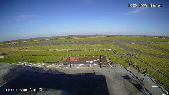A wide view of an airport runway taken from a high vantage point. In the foreground, there is a helipad marked with a large white "X" on a dark surface. Surrounding the helipad is a grassy area with a white fence. Beyond the helipad, a runway stretches across the image, bordered by a green field on both sides. There are no aircraft visible, and the sky is clear with a few wispy clouds. The temperature reading in the corner indicates the time as 14:59:52 on March 26, 2025.
