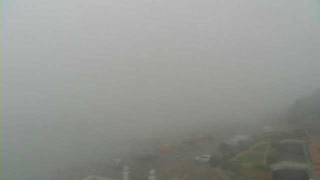 A foggy scene showing a nearly obscured coastal area. The view is mostly gray with limited visibility, making it difficult to see details. In the foreground, there are faint outlines of vehicles parked along a road, and hints of shapes suggesting buildings or structures nearby. The ocean is visible but shrouded in mist, blending into the foggy atmosphere.