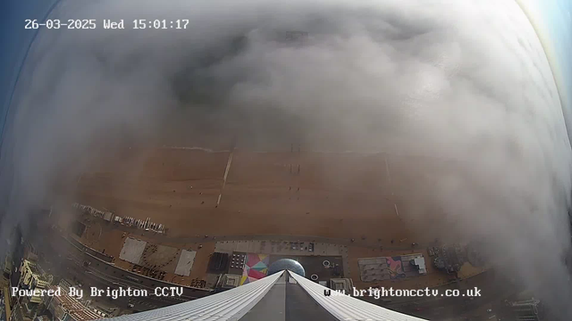A view looking down from a high vantage point showing a sandy beach covered by a light fog. The beach is lined with various structures, including a colorful playground area, and there are a few small figures in the distance. The scene is partially obscured by cloud cover, creating a soft, muted atmosphere. The image includes a timestamp indicating the date and time, as well as a watermark for Brighton CCTV at the bottom.