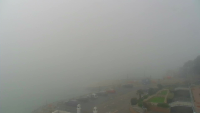 A foggy seaside view with very low visibility. The scene appears gray and muted, obscuring details of the water and shoreline. In the foreground, there are outlines of parked cars along a road, while faint shapes of green grass and stone steps can be seen nearby. The details of buildings or structures are mostly indistinct due to the thick fog.