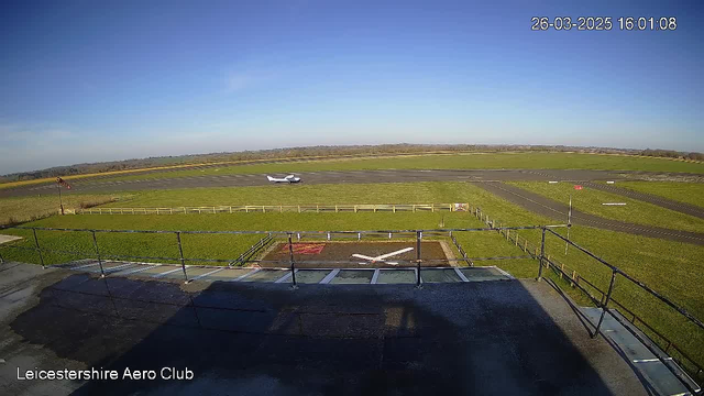 A clear blue sky is visible above a grassy airfield. A small aircraft is taxiing on the runway, which is marked with a few lines and signs. In the foreground, a fenced area with a white "X" marking is present, alongside a railing that suggests a viewing platform. The landscape extends towards a distant horizon with faint rolling hills.