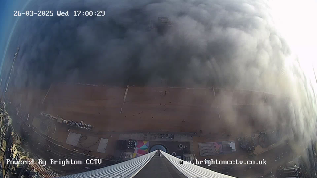 A wide-angle view from a high vantage point looking downwards, capturing a beach and a boardwalk. The image features a foggy sky, obscuring visibility of distant structures. The beach below has a sandy area with people walking, and colorful beach huts or stalls are visible along the promenade. The time displayed at the top right indicates it is 17:00 on March 26, 2025. The bottom left corner displays the text "Powered By Brighton CCTV" with the website link below it.
