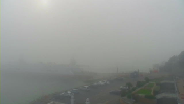 The image shows a waterfront scene largely obscured by thick fog. In the foreground, there is a parking lot with several cars visible. To the right, there are patches of green grass and garden areas. The water is also visible but appears dim due to the fog, and a faint outline of a ship can be seen in the distance. The background features blurred silhouettes of buildings and trees, with a bright light, likely the sun, faintly shining through the fog.