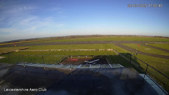 A wide view of an airfield from a raised position, featuring a clear blue sky with a few clouds. In the foreground, there is a metal railing, and the ground consists of green grass and gray asphalt. A faint white cross marking is visible on the ground. The airfield is bordered by a white fence. In the distance, fields and trees can be seen, indicating a rural setting. A small red flag is positioned to the right of the image, indicating the wind direction. The bottom left corner displays the text "Leicestershire Aero Club" along with a timestamp in the top right.