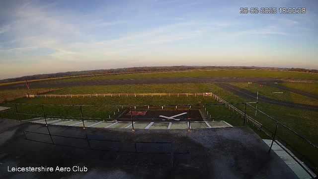A view of a runway at Leicestershire Aero Club during the late afternoon. The sky is mostly clear with a few clouds, and the sun is low, casting a warm light over the surroundings. In the foreground, a helipad with a red and white numbering and a white cross symbol is visible, surrounded by a railing. The runway extends into the distance, lined with green grass on either side, and more runway markings in the background. The scene is tranquil, showcasing an open expanse of land.