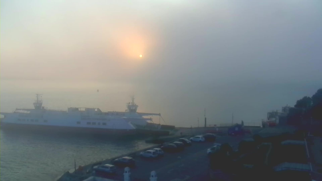 A foggy seascape at dawn, with a muted sun rising in the background. In the foreground, a large ferry is docked near the water's edge, and there are several cars parked along the waterfront. The scene is shrouded in mist, creating a soft, ethereal atmosphere. The water appears calm, with faint outlines of distant objects barely visible through the haze.