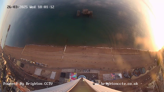 A high-angle view of a beach with golden sand, gentle waves lapping at the shore, and a calm blue sea. There is an old pier extending into the water and a visible jetty. In the foreground, various beach facilities and people can be seen walking along the shore. The image is taken during sunset, casting a warm, soft light over the scene. The date and time are displayed in the upper left corner.