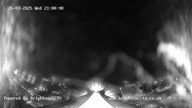 A black-and-white webcam image shows a partially blurred view of a narrow, bright pathway leading into darkness. On the left side, faint outlines of buildings are visible, with lights reflected on a wet surface. Text at the bottom indicates the image is powered by Brighton CCTV and the time and date displayed in the upper corner is 21:00:48 on March 26, 2025.