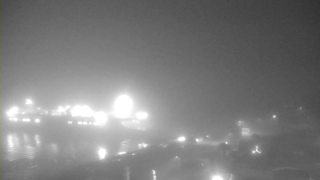 A foggy waterfront scene at night with limited visibility. Blurry silhouettes of boats and structures are faintly visible. Bright lights from a nearby dock or ship create circular glows in the mist, reflecting off the water's surface. The setting appears quiet and eerie due to the heavy fog.