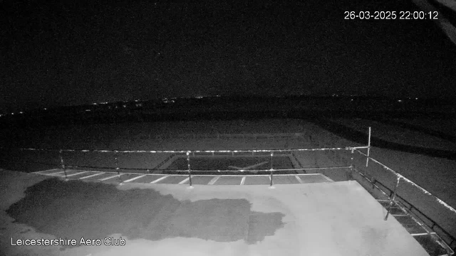 A dark image taken from a webcam at night, showing a flat surface that appears to be a runway or an airfield. There is a railing or fence along the edge of the surface. In the background, faint lights from distant buildings or vehicles are visible. The image has a timestamp indicating it was taken on March 26, 2025, at 10:00 PM.
