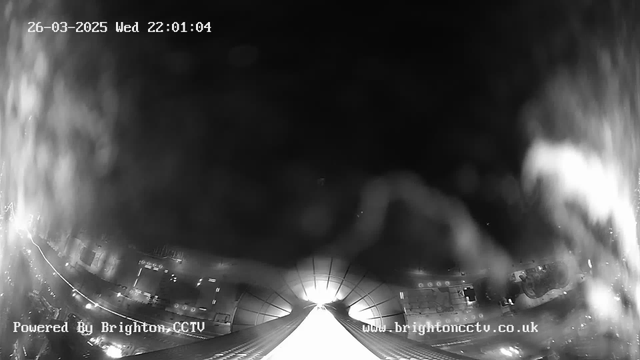 A black and white image from a webcam showing a view down a brightly lit street at night. The bottom half of the image features a lighted pathway leading off into the distance, possibly a roadway or walkway, while the upper half shows a dark sky with some blurriness, suggesting a wet lens. There are faint outlines of buildings and scattered lights on the sides of the street. The date and time are displayed in the top left corner. The image has a slight distortion, adding a watery effect.