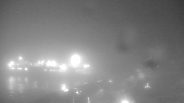 A heavily foggy scene showing faint outlines of a waterfront area. Bright lights from boats or buildings are partially visible through the fog, creating a blurred effect. The water reflects some of the lights, adding to the hazy atmosphere. Overall, visibility is low, with a monochromatic color scheme due to the fog.