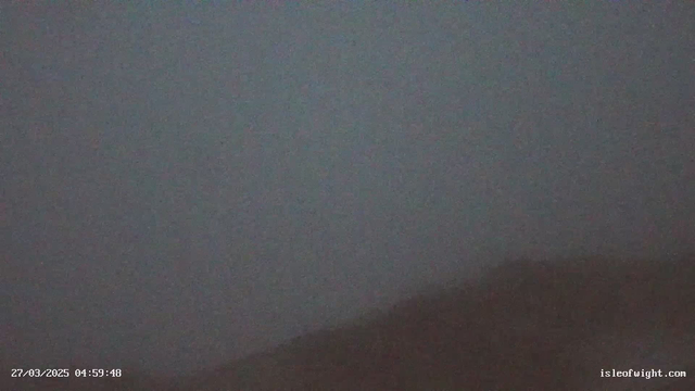 A foggy landscape with very low visibility, primarily showing muted shades of gray. The upper part of the image appears slightly lighter, while the lower part features indistinct shapes that suggest a dark, uneven terrain. The date and time are displayed in the lower left corner, indicating early morning.