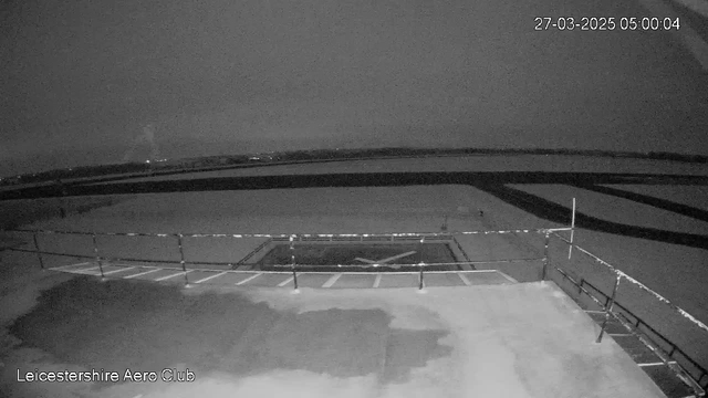 A dimly lit scene showing a flat surface with a helipad outline in the center. There is a railing in the foreground on the left side, and the background features a cloudy sky above a dark landscape with faint lights in the distance. The timestamp reads 27 March 2025, 5:00 AM.
