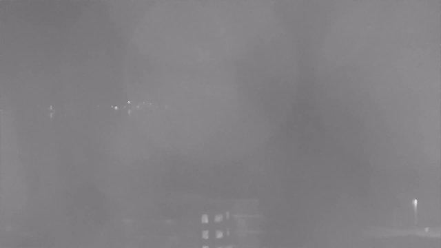 A dimly lit scene showing blurred outlines of buildings and distant lights, with a significant amount of haze or fog obscuring visibility. The overall image appears gray and indistinct, suggesting nighttime conditions.