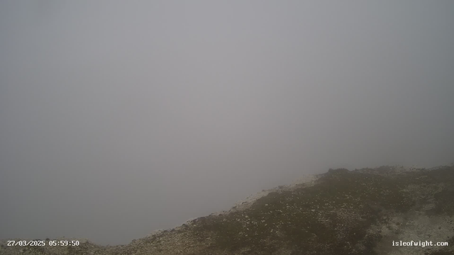 A foggy landscape is depicted, with a gray sky blending into a dense mist. The view is obscured, making it difficult to see any distinct features in the distance. In the foreground, there is a patch of ground that appears to have some grass and rocks, but the visibility is very limited overall due to the fog. The scene conveys a sense of stillness and muted colors.