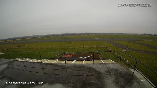A view from a webcam at Leicestershire Aero Club shows an airport runway and surrounding grassy field. The sky is overcast and gray, indicating a cloudy day. In the foreground, there is a rooftop railing with a section of the ground marked for landing with a white cross shape and a red area nearby. The runway extends into the distance, with a small wind sock visible to the left, indicating wind direction.