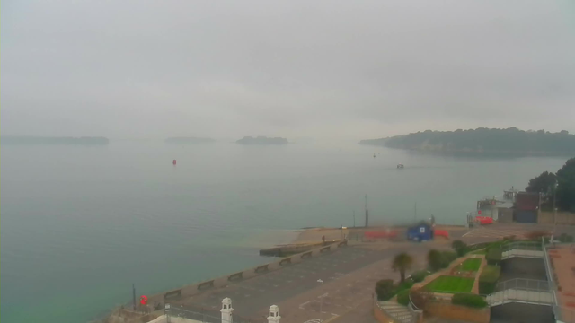 A foggy view of a calm body of water with distant islands partially obscured by mist. In the foreground, a paved area leads down to the water, with a few small structures and greenery visible. A red buoy is seen in the water, and a small boat is traversing the scene. The overall ambiance is tranquil and muted due to the cloud cover.