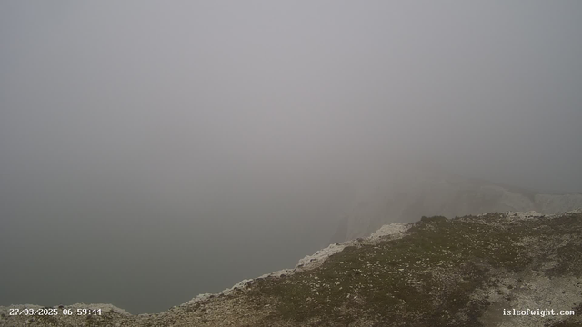 A foggy landscape where visibility is extremely low. The image shows a rocky cliff edge at the right side, with a grassy area in the foreground. The background fades into a thick mist, obscuring any distant features, creating a serene yet unclear atmosphere. The timestamp at the bottom indicates the date and time.