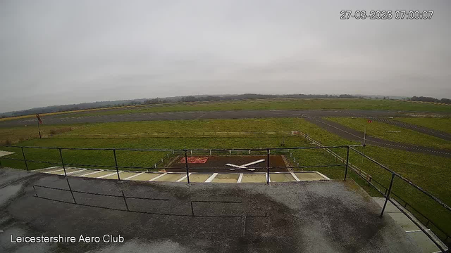 A gray, overcast sky is visible above a grassy airfield. In the foreground, there is a railing along the edge of a platform, overlooking a helipad marked with a white "X." The airfield features a paved runway with patches of grass around it, and there's a wind sock visible on a pole. The scene appears calm and empty, with no aircraft in sight. The date and time at the top indicate March 27, 2025, at 07:00.