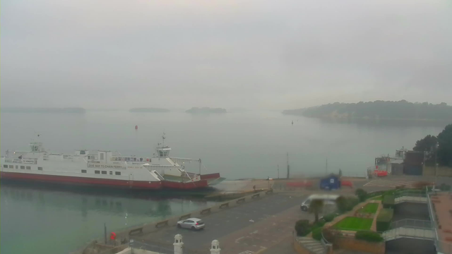 A misty waterfront scene featuring a large ferry docked at a pier. The ferry has a white and red color scheme. The water is calm and reflects the overcast sky, with distant islands visible in the background. A small, parked car is on the road near the ferry terminal, and there are patches of greenery and stone pathways along the waterfront. The overall atmosphere is serene and slightly foggy.