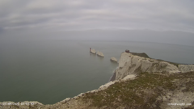 A cloudy sky stretches over a calm sea, with soft waves lapping against a rocky coastline. In the foreground, grassy cliffs are visible, leading down to the water. An outcrop of white chalk cliffs juts out into the sea, with a small, red lighthouse positioned on one of the rocks. In the background, additional rugged cliffs can be seen, disappearing into the misty horizon. The atmosphere is serene and somewhat overcast, with muted colors dominating the scene.