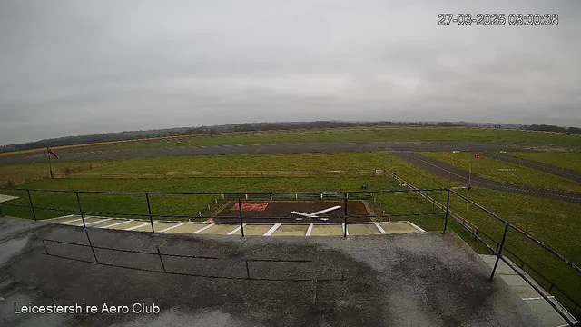 A cloudy sky is visible above an open field, with a runway stretching across the middle of the scene. On the left side, there is a red and white windsock indicating wind direction. In the foreground, a helipad marked with a white cross is seen on a dark surface. Fencing surrounds parts of the area, and the background shows rolling hills in the distance. The image is timestamped at 08:00:38 on March 27, 2025.