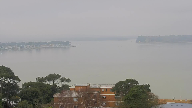 A cloudy view of a calm body of water, with a shoreline and several boats visible in the distance. In the foreground, there are brown buildings partially obscured by trees. The atmosphere appears misty and overcast. The horizon blends with the water, creating a seamless transition between the sky and the lake.